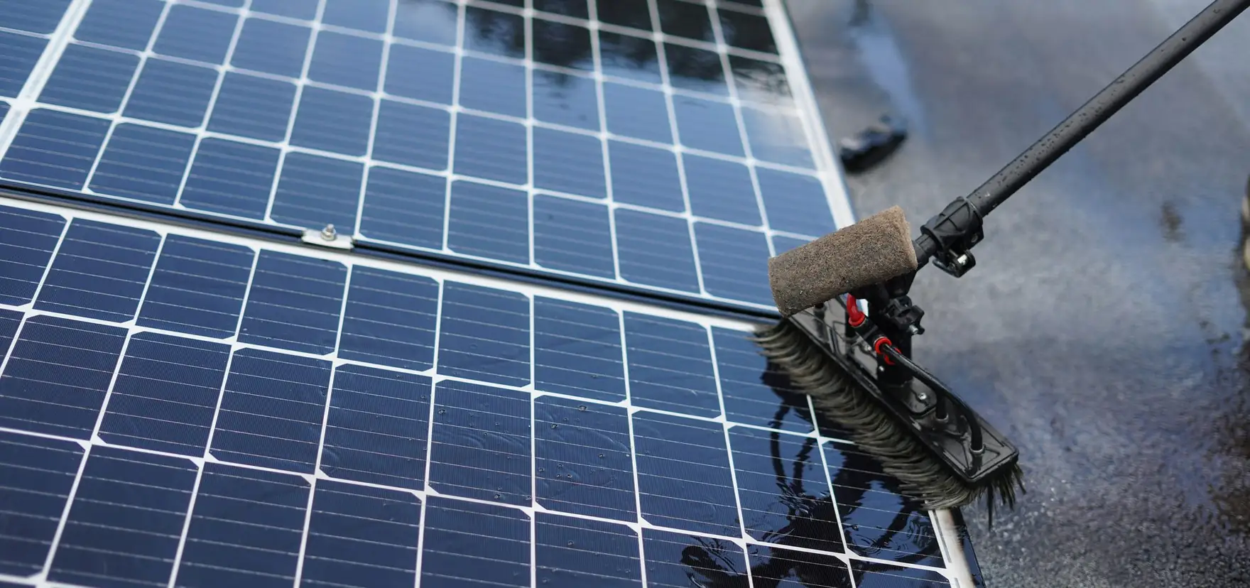 A technician cleaning a rooftop solar panel with a wet brush on a telescopic pole